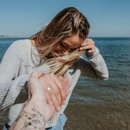 Person with the tattoo, holding hand of the beautiful, blonde woman, near the water