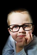 Portrait of the child boy with glasses, holding head on hand, in light, at dark background