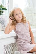 Cute, blonde girl with curly hair, in dress, posing near the windows with light