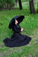 Woman in black dress, and with black umbrella, posing among the green grass and trees