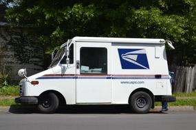 Mail truck, on the road, in sunlight, near the colorful plants and fence