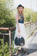 Blonde woman with curly hair, in dress, hat and with the bag, posing near the bridge, among the colorful plants