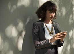 Woman with curly hair, in suit, using smartphone near the fence in light and shadows