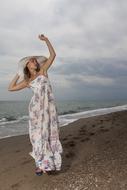 Beautiful woman in colorful dress and hat, posing on the beach, under the clouds