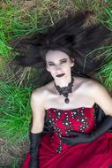 Gothic woman with make-up, and in the red, black and white dress, laying on the green grass