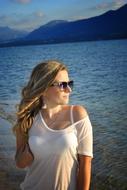 Blonde woman in white clothing, posing on the beautiful shore with mountains