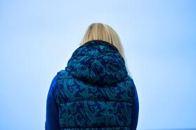 Back view of the blonde woman in blue coat and in blue, patterned vest, at blue sky background