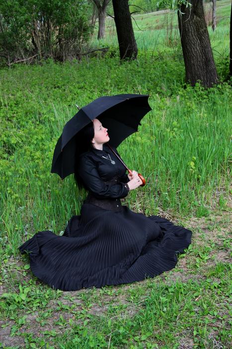 Woman in black dress, and with black umbrella, posing among the green grass and trees