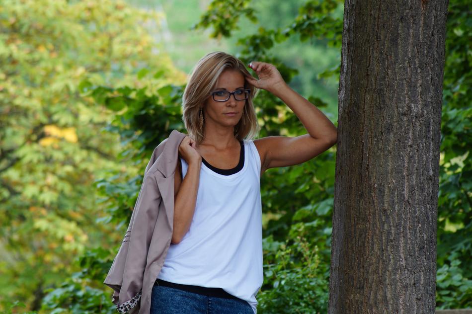Woman in glasses and with beige jacket posing near the tree in the beautiful and colorful forest