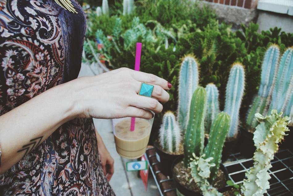 Woman with the cup of coffee with purple straw, standing near the beautiful cactus plants