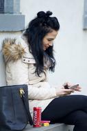 Young Woman in winter clothing sits with smartphone at wall