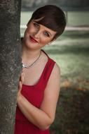 young Woman in Red Dress posing behind tree trunk