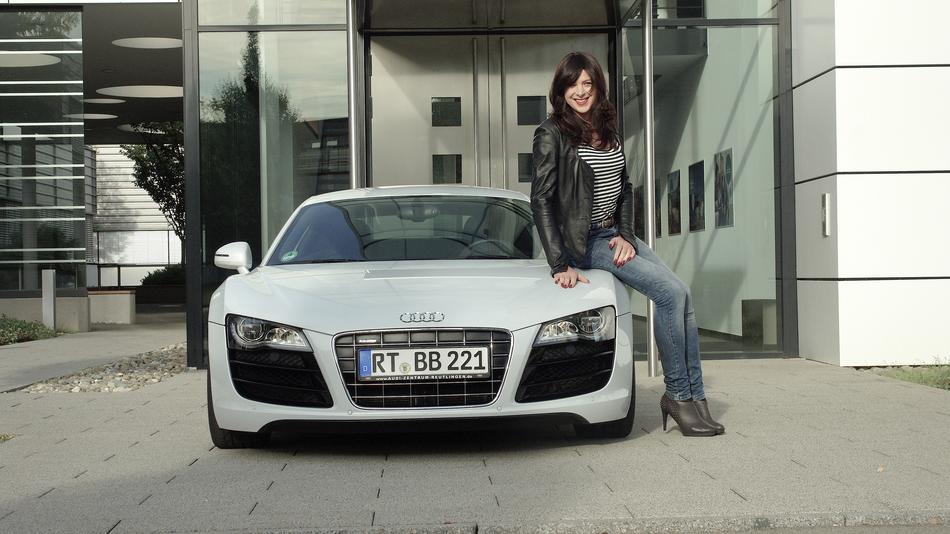 Smiling woman posing on the grey 'Audi R8' car near the building