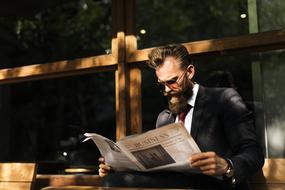 brutal man with a beard is reading a newspaper while sitting on a bench