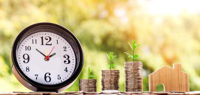 Clock time and shiny, growing money coins and wooden home, at blurred background with colorful plants