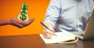 light bulb with dollar sign inside on hand beside of person with laptop at desk