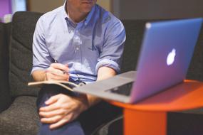 working man with pen and notebook sits on sofa in front of laptop