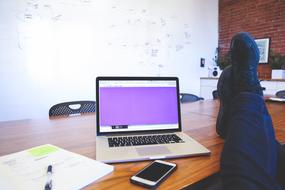 Person, sitting with the legs on the wooden desk, with the Mackbook laptop, Iphone smartphone, papers and pen