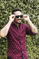Smiling, bearded man, with the tattoos, holding sunglasses, near the colorful plants