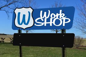 Blue and white "Work Shop" street sign with logo, among the colorful fields and plants