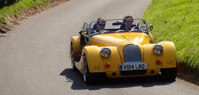 People driving shiny, yellow "Morgan" electric car, on the road in sunlight, among the green grass