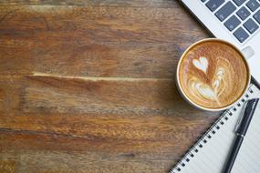 Cup of coffee, with the beautiful drawing, near the notebook with the pen, near the laptop, on the wooden surface