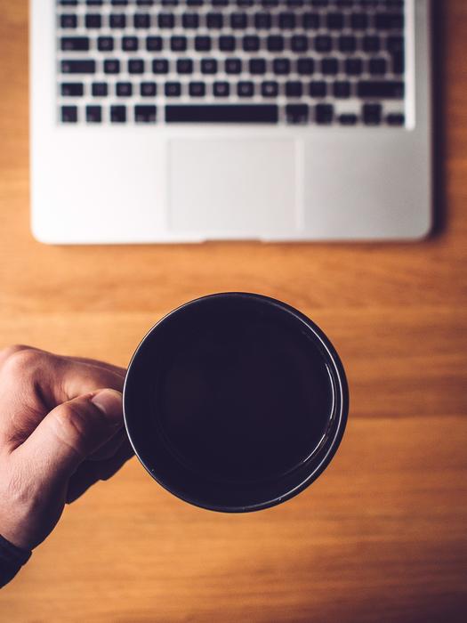 Person, holding cup of coffee, above the wooden desk, with the laptop, in light