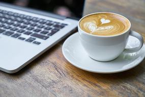 coffee wit heart shape art in white cup beside of Laptop Computer