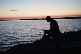 Man sits with phone on coast at dusk