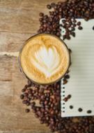 Beautiful cup of coffee with the beautiful heart drawing, among the brown coffee beans and notebook on the wooden table