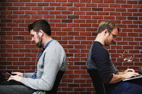 Sitting men, using devices, near the brick wall