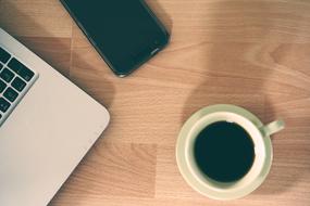 Cup of the beautiful coffee, near the black smartphone and laptop, on the wooden surface