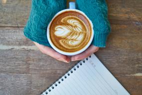 Person in turquoise sweater, holding a cup of coffee with the beautiful drawing, near the notebook, on the wooden table