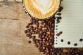 Beautiful cup of coffee with the drawing, near the notebook, among the brown coffee beans, on the wooden surface