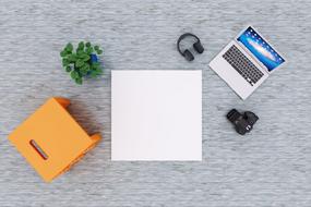 White notebook, near the orange seat, green plant, camera, headphones and laptop