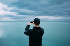 Back view of the guy, taking photo of the beautiful, turquoise sea, under the clouds