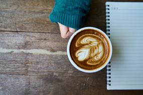 Cup of coffee, with the beautiful drawing, near the hand in turquoise sweater, near the notebook, on the wooden table