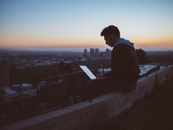 young man sits with laptop computer above city at dusk