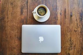 Silver Macbook with white logo and white cup of coffee on white plate on the wooden surface