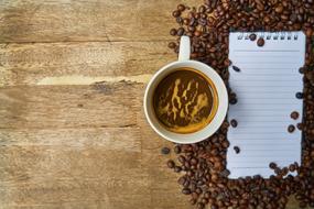 Cup of the beautiful coffee, near the brown coffee beans and notepad, on the wooden surface