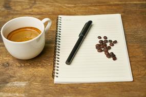 Cup of coffee, near the notebook with the pen and brown coffee beans, on the wooden surface