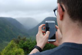 Person with glasses, taking photo of the beautiful green mountains in white clouds, with smartphone