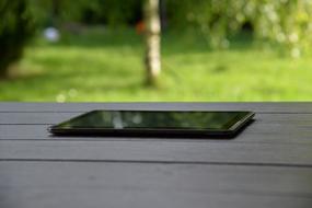 Close-up of the shiny, black tablet, on the wooden table, in shadow, among the beautiful, green plants