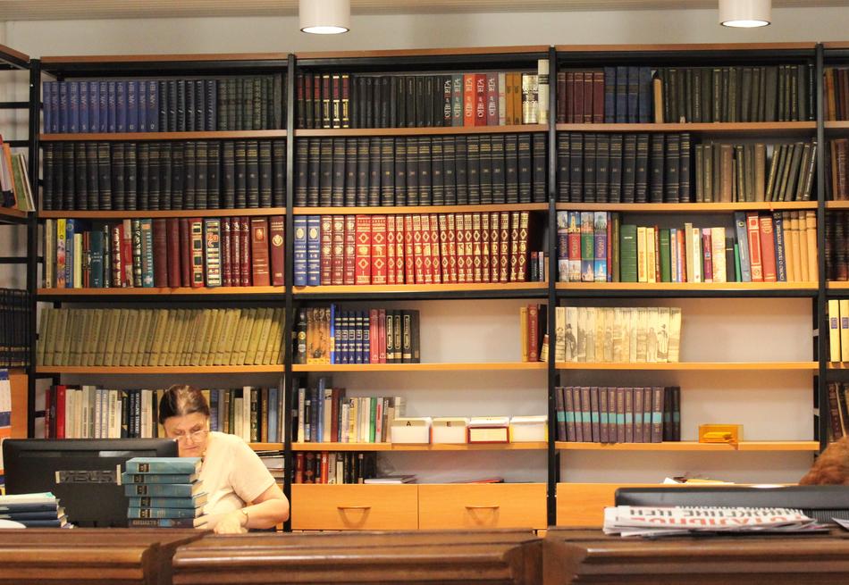 Senior woman works at desk in light in front of Rack with Books