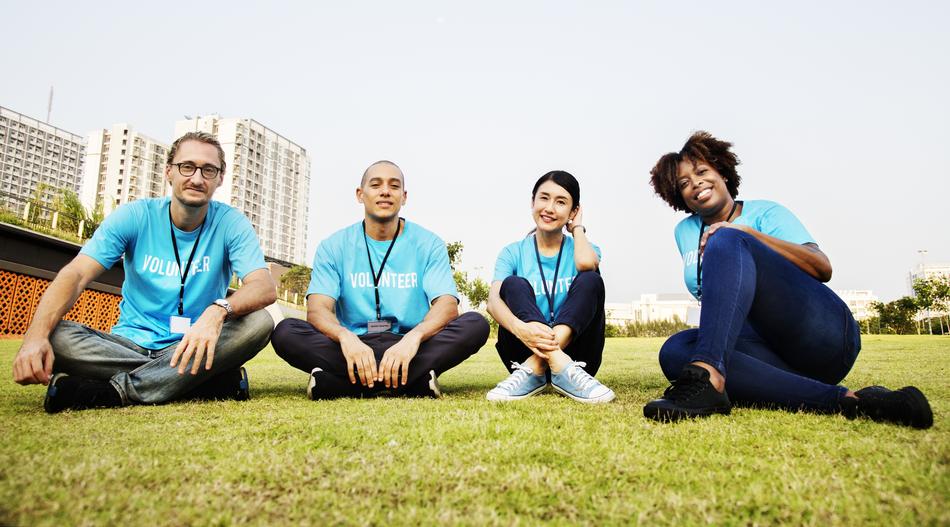 Smiling volunteers, sitting on the green meadow, near the buildings