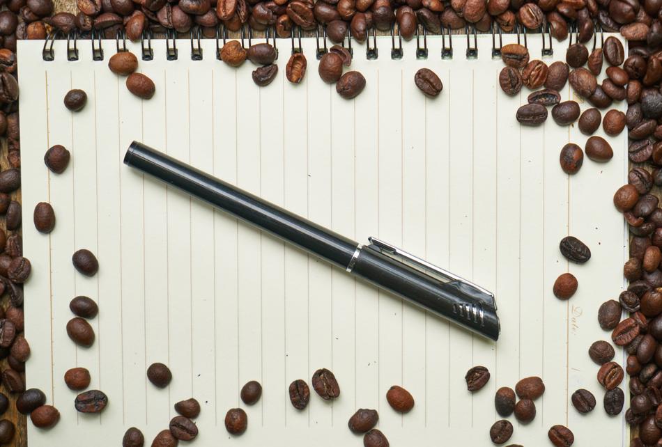 Close-up of the shiny pen, on the notebook, with the lines, among the beautiful, brown coffee beans