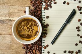 Cup of the beautiful coffee, near the notebook with pen, in coffee beans, on the wooden surface