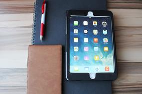 Ipad with the colorful icons, in black case, near the brown notebook and pen, on the black notebook, on the wooden surface