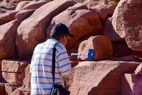 Man in cap, with the bag and smartphone, near the rocks in sunlight