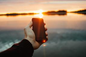 Person holding smartphone above the water, at colorful sunset